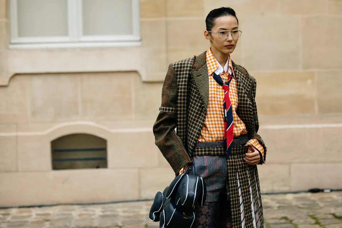 Google AI, Universal Commerce Protocol i Business Agent. Stylowa osoba w okularach spaceruje ulicą, odziana w warstwową stylizację z kraciastym okryciem wierzchnim, pomarańczową koszulą w kratę, musznikiem i wzorzystymi spodniami, dzierżąc czarną torbę na tle jasnej kamiennej fasady.