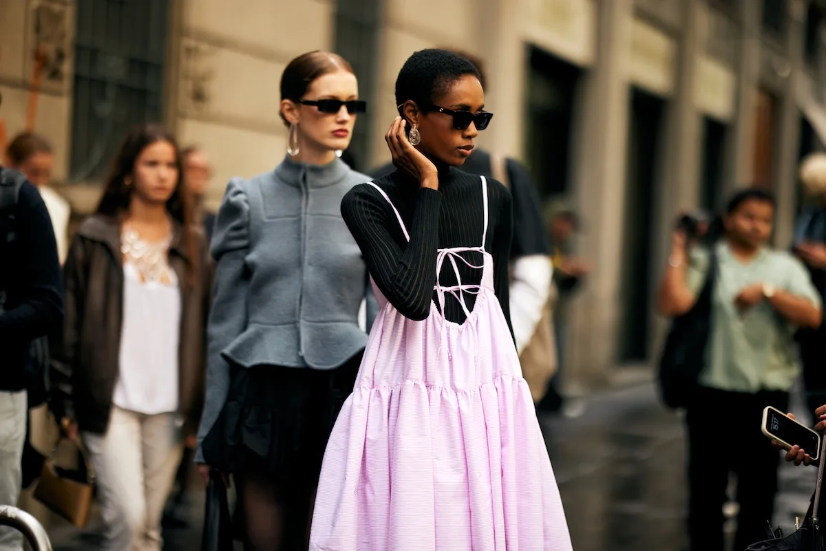 Stylowe uczestniczki street style w czasie Fashion Week w Mediolanie – kobieta w czarnym swetrze z golfem i bladoróżowej sukience na ramiączkach oraz modelka w popielatej marynarce i ciemnych okularach przeciwsłonecznych, sfotografowane w miejskim otoczeniu.