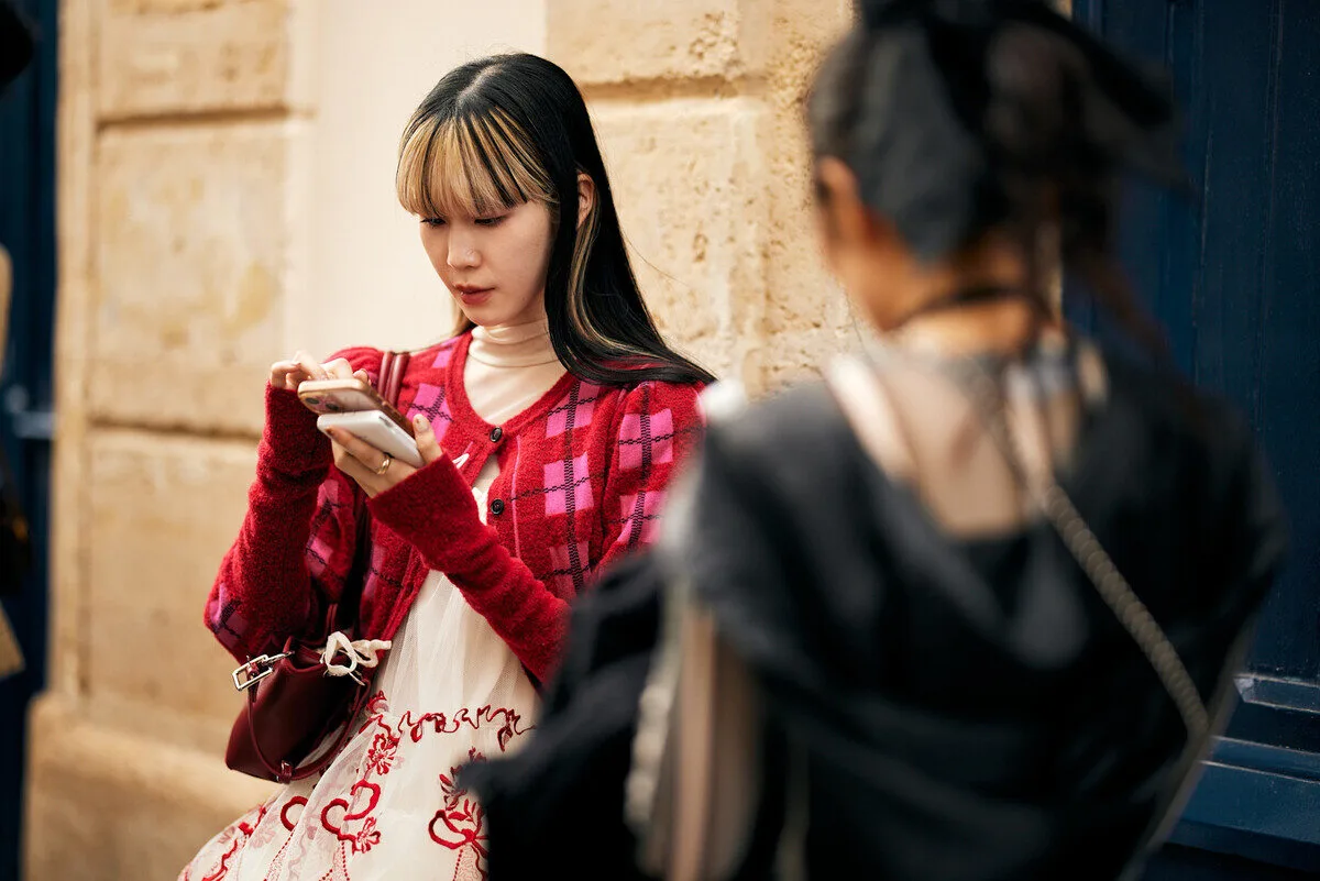 Zakaz mediów społecznościowych dla dzieci w Polsce od 2026? KO szykuje projekt ustawy. Młoda kobieta z dwukolorowymi włosami i w czerwonym, kraciastym kardiganie skupiona na ekranie smartfona, stojąca na tle piaskowej ściany budynku.