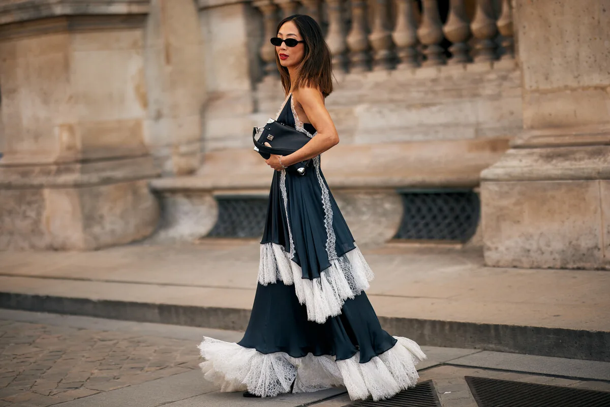 Open AI przejęcie Pinterest. Na zdjęciu: stylizacja street style z Paryża: kobieta w czarnej, warstwowej sukni z białymi koronkowymi falbanami, w okularach przeciwsłonecznych, trzymająca czarną kopertówkę na tle kamiennej architektury.