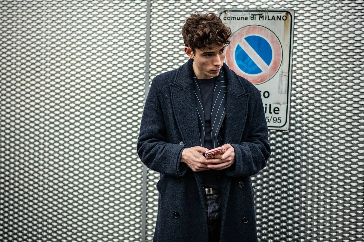 Ogłoszenia w ChatGPT. Mężczyzna odziany w szykowny, ciemny płaszcz i marynarkę w paski stoi obok metalowej bariery, z uwagą obserwując wyświetlacz swojego smartfona. Jego koncentracja znakomicie ilustruje moment, kiedy użytkownik natrafia na reklamy w ChatGPT, skrojone do jego indywidualnych upodobań. Ten obraz symbolizuje połączenie klasycznej elegancji z nowoczesną, algorytmiczną sferą marketingu online.