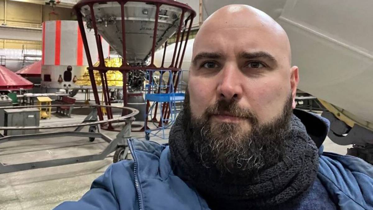 Mężczyzna z brodą w niebieskiej kurtce i szaliku na tle konstrukcji przemysłowej.