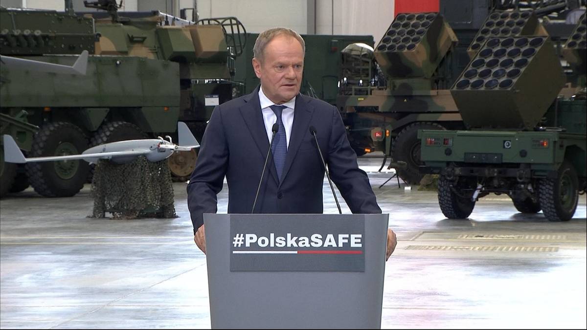 Premier Donald Tusk przemawia przy mównicy z napisem "#PolskaSAFE", w tle widoczne wozy bojowe i dron.