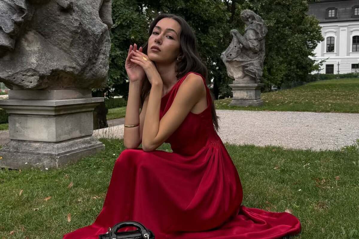 Ukraińska marka Vovk wycofuje się z Polski. Kobieta w długiej, czerwonej sukni siedzi na trawie w otoczeniu kamiennych rzeźb i zieleni, z elegancką czarną torebką u boku.