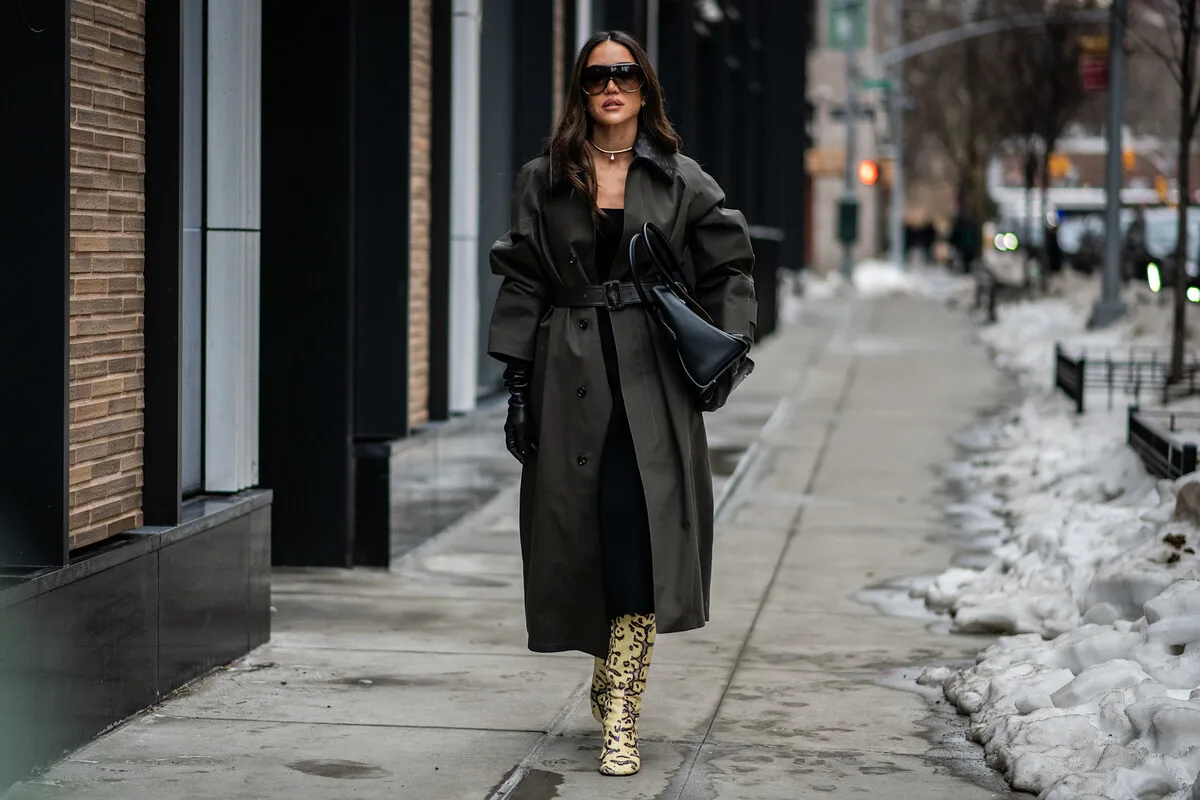 Fałszywe kontenery PCK Warszawa. Kobieta idąca chodnikiem w miejskiej scenerii zimą, ubrana w długi oliwkowy płaszcz z paskiem, czarne rękawiczki, duże okulary przeciwsłoneczne i trzymająca czarną torebkę; na nogach ma wzorzyste, wysokie kozaki w panterkę.