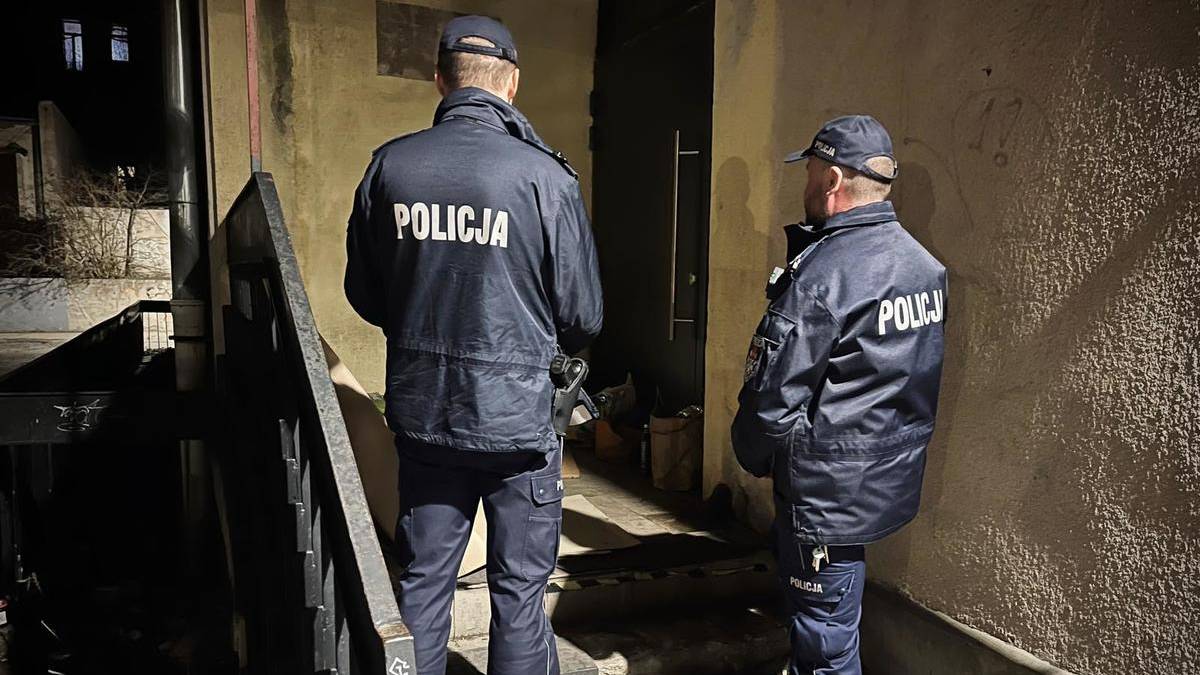 Dw&oacute;ch policjant&oacute;w w kurtkach z napisem "POLICJA" stoi na zewnątrz budynku