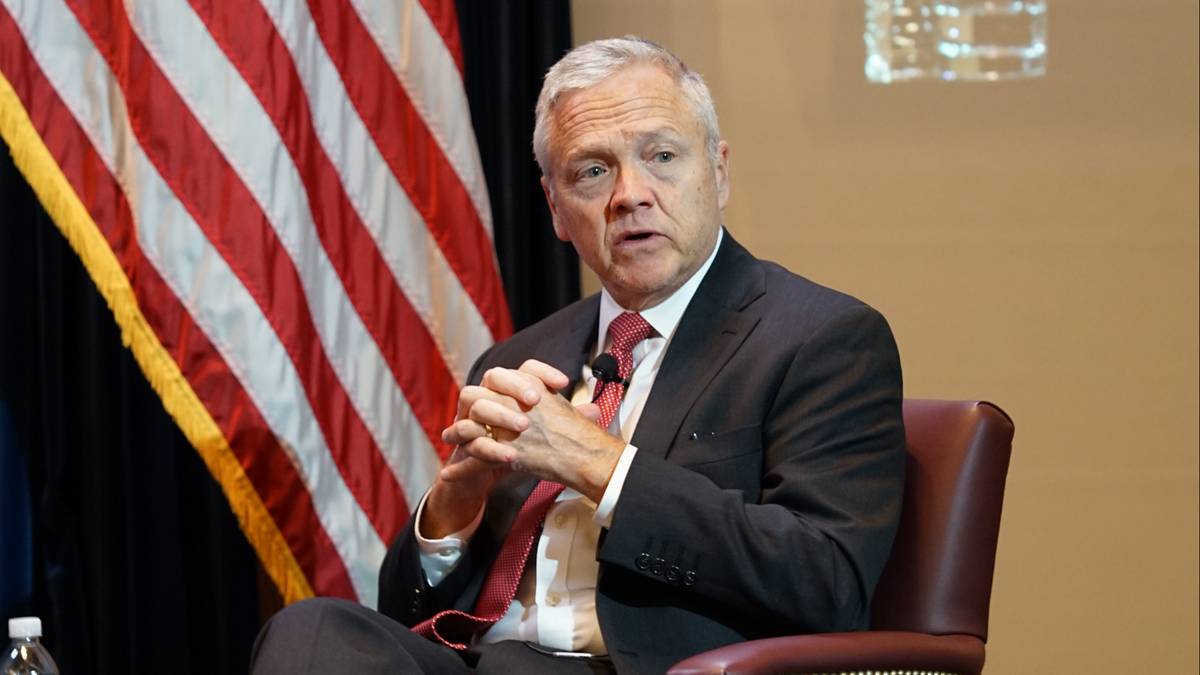 Mężczyzna w garniturze i krawacie siedzi na krześle, z amerykańską flagą w tle.