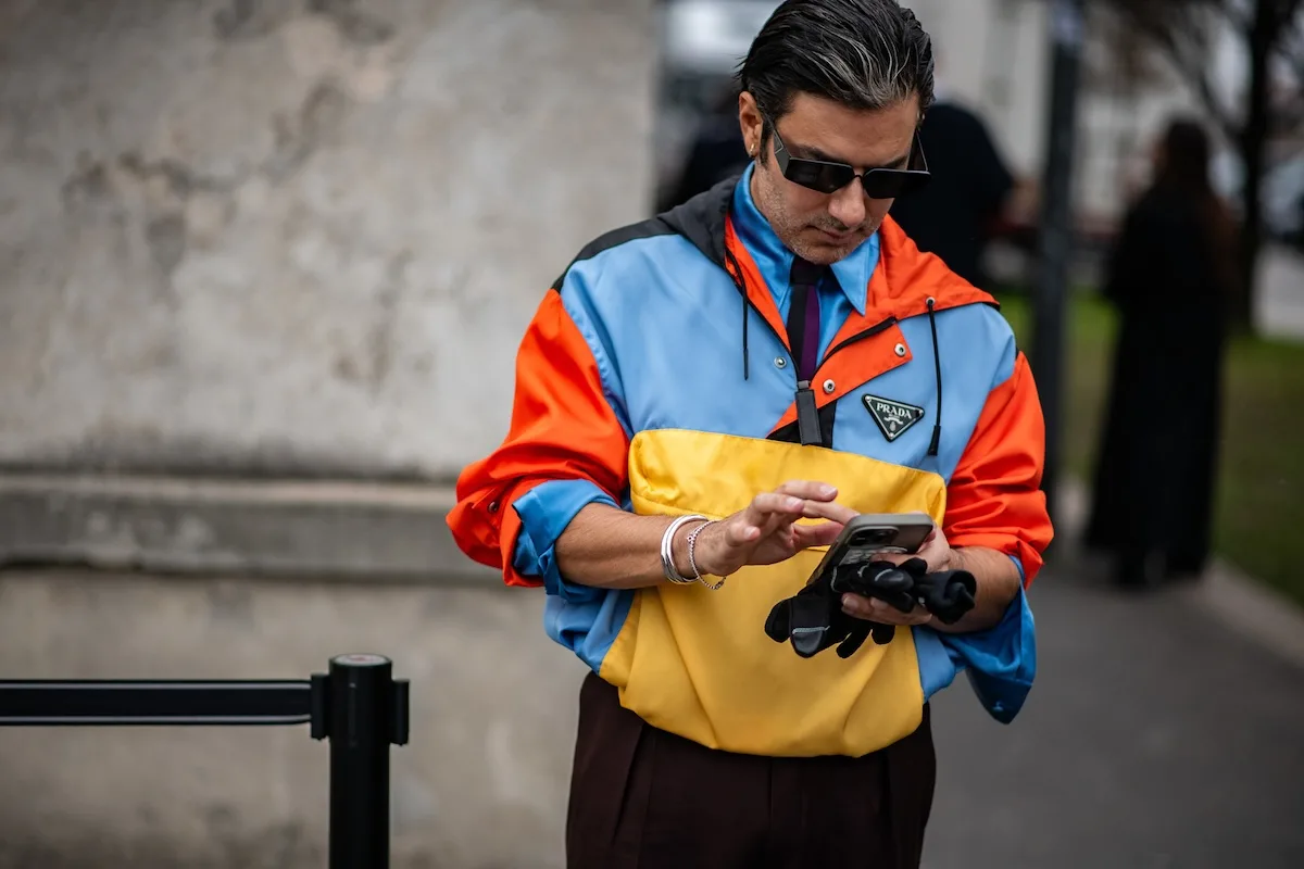 Mężczyzna w kolorowej kurtce Prada typu color block sprawdzający telefon podczas street style na pokazach mody w Mediolanie.