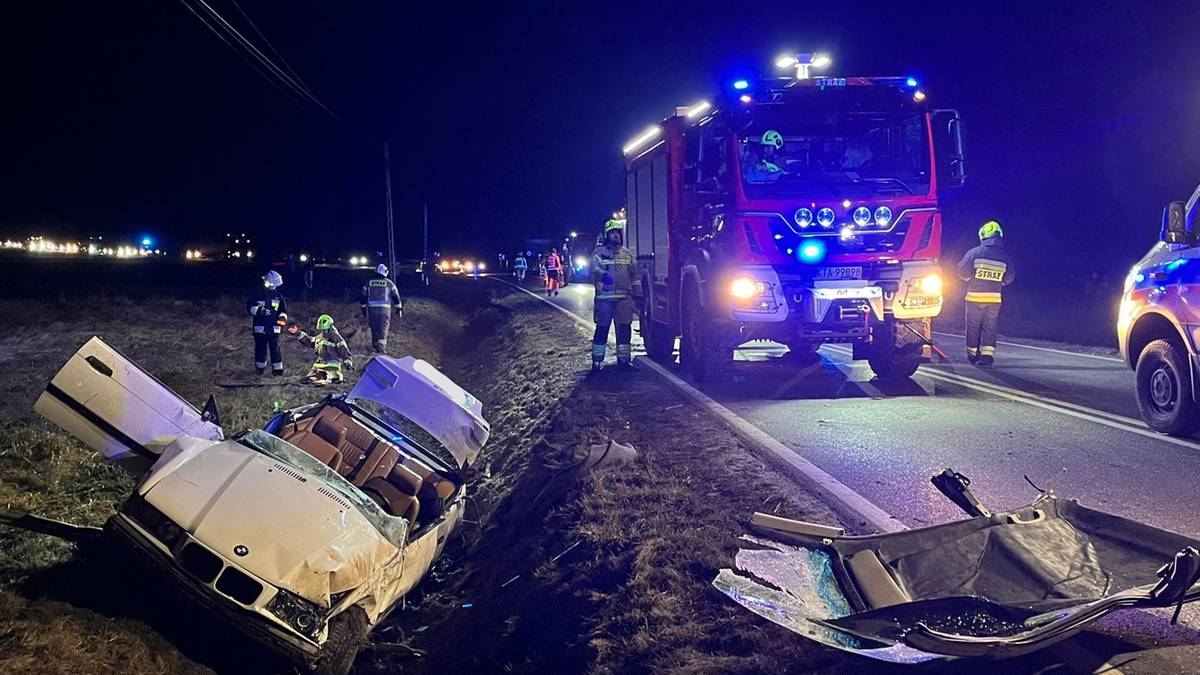 Samoch&oacute;d po wypadku drogowym w nocy, w&oacute;z strażacki i ratownicy na poboczu