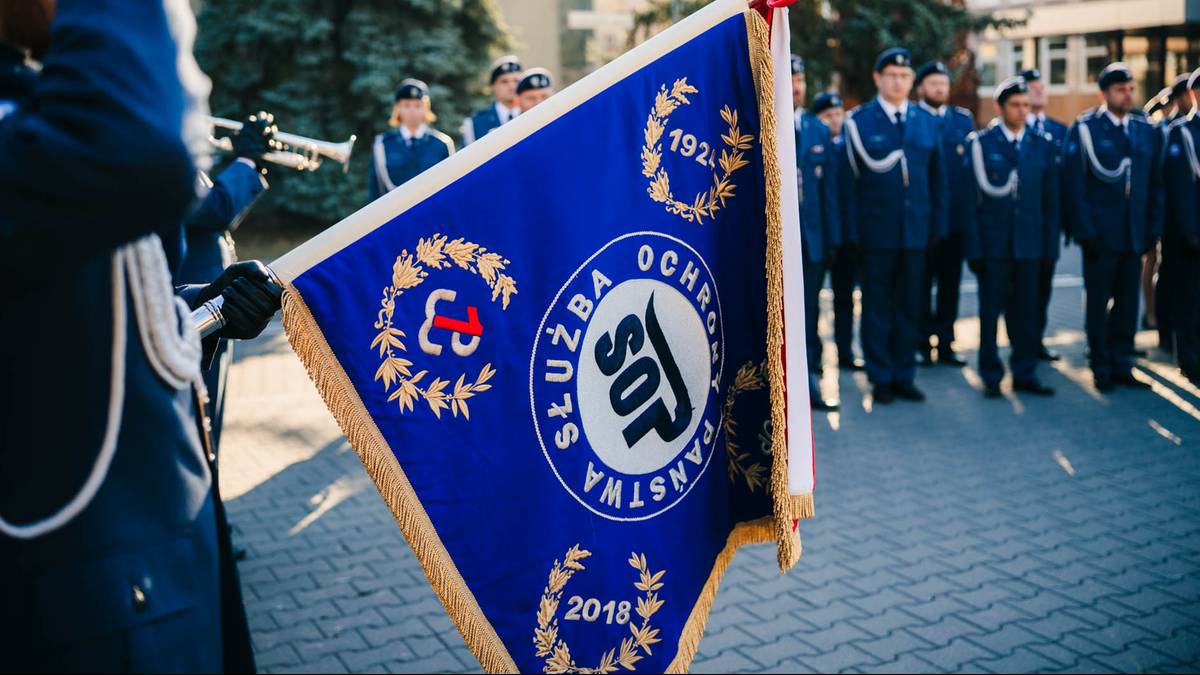 Fragment flagi Służby Ochrony Państwa z widocznym herbem i rokiem 2018, w tle stojący w szeregu funkcjonariusze formacji.
