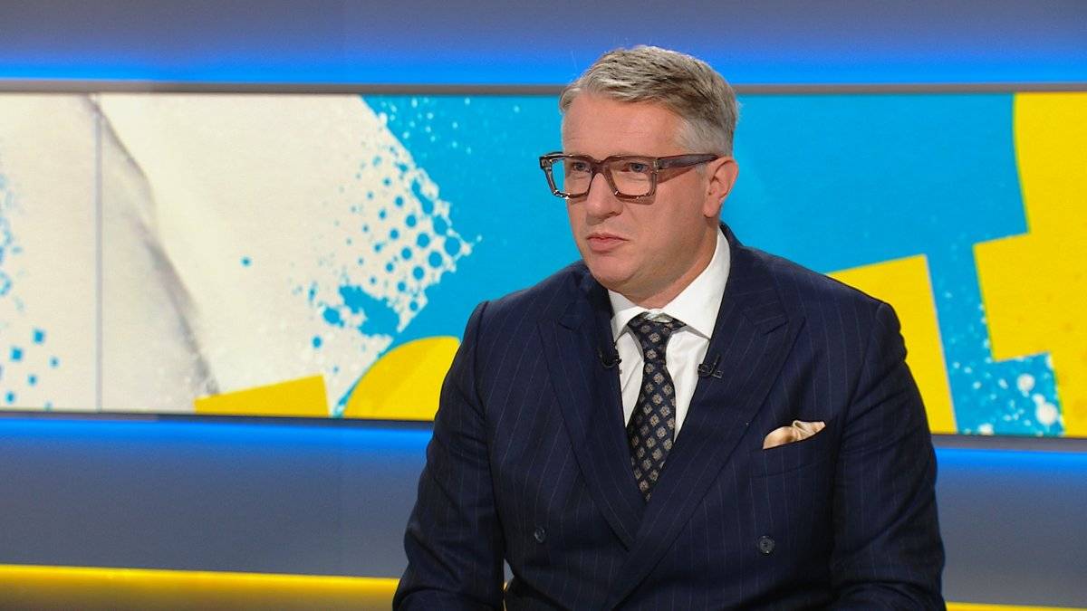 Mężczyzna w okularach i granatowym garniturze siedzi w studiu telewizyjnym.