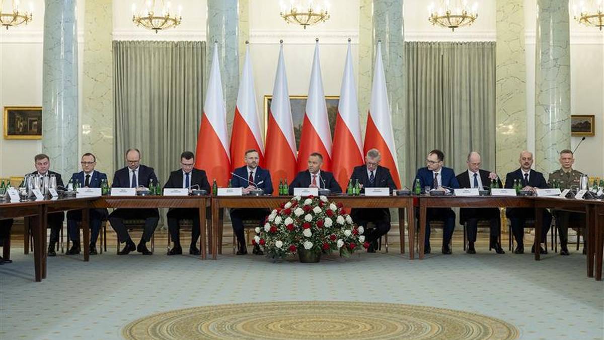 Grupa mężczyzn w garniturach siedzi przy stole pod polskimi flagami, podczas spotkania Rady Bezpieczeństwa Narodowego.