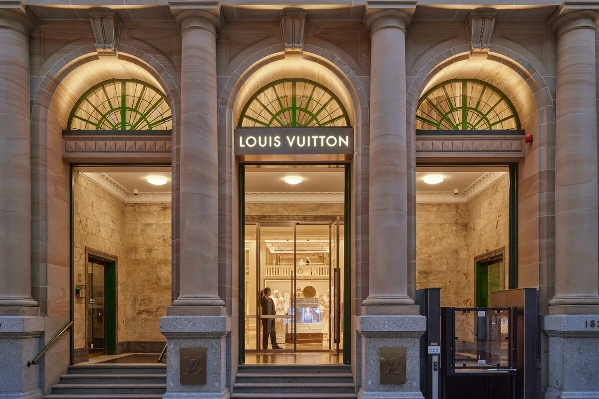Louis Vuitton kara pranie pieniędzy. Wejście do butiku Louis Vuitton w eleganckiej, kamiennej kamienicy z wysokimi kolumnami i łukowymi oknami; nad przeszklonymi drzwiami widoczny podświetlany szyld marki, a wewnątrz jasne, luksusowe wnętrze z ekspozycją torebek.