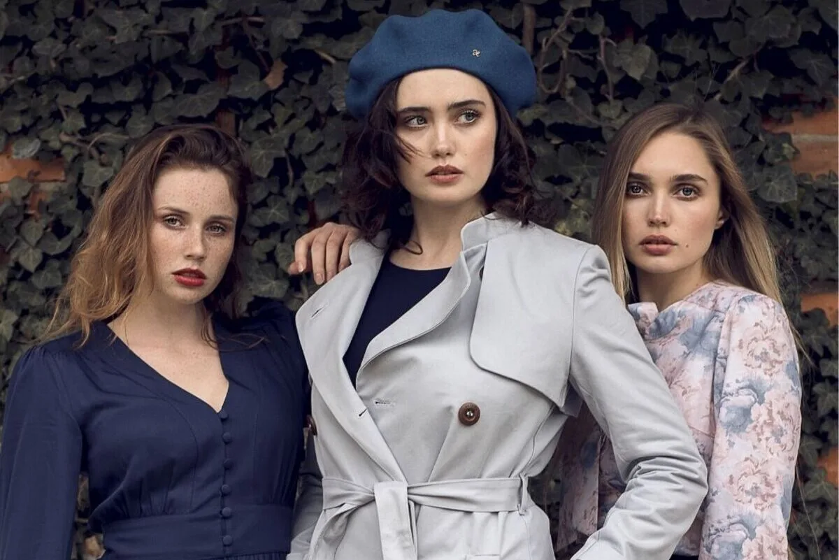 Aeterie skargi, nowa strategia funkcjonowania. Trzy modelki w szykownych, ponadczasowych kreacjach inspirowanych stylem retro, pozujące na tle zielonej ściany z roślinami pnącymi.