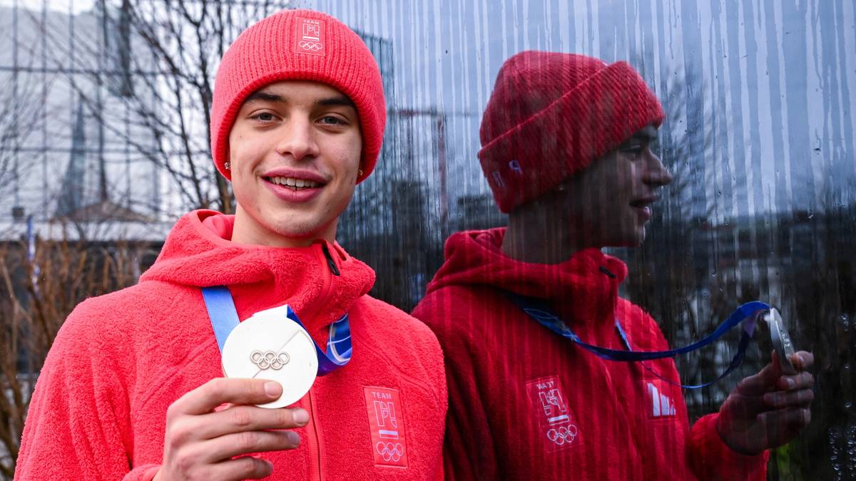 Młody mężczyzna w czerwonej czapce i bluzie z kapturem, trzyma w dłoni srebrny medal olimpijski.