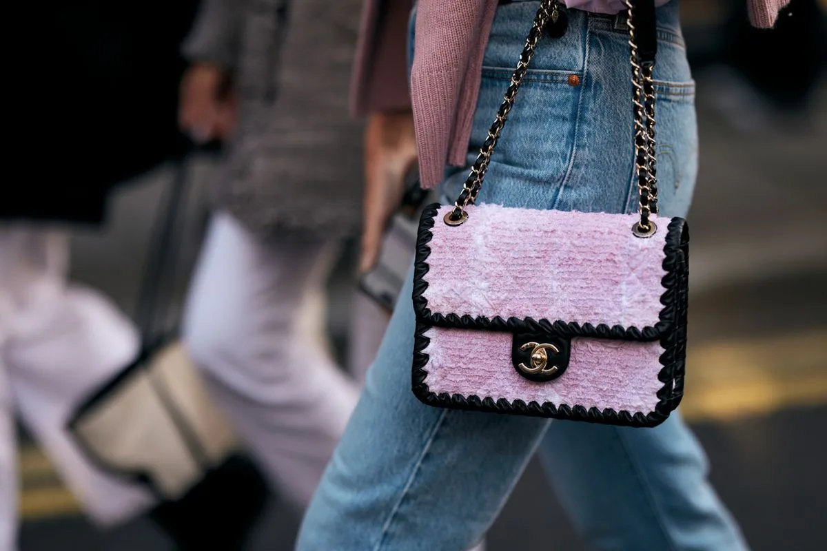 Fotografia prezentuje stylizację uliczną z Tygodnia Mody w Londynie, ukazując kobietę w niebieskich dżinsach z wysokim stanem i jasnoróżowym pulowerze. Kluczowym elementem kompozycji jest ekskluzywna torebka Chanel, uszyta z różowej, teksturowanej tkaniny typu tweed z czarnym wykończeniem i złotym zamknięciem. Ten szczegół podkreśla wysoką jakość dodatków premium, na które mogą mieć wpływ świeże przepisy podatkowe w sektorze mody. Tło jest delikatnie zamazane, eksponując dynamiczny ruch miejski i inne osoby o modnym wyglądzie.