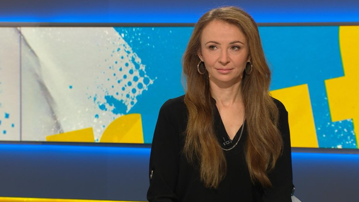 Kobieta w czarnej bluzce z dekoltem w kształcie litery V, z długimi, falowanymi blond włosami, spogląda w kierunku kamery. Tłem dla niej jest niebiesko-żółty deseń graficzny.