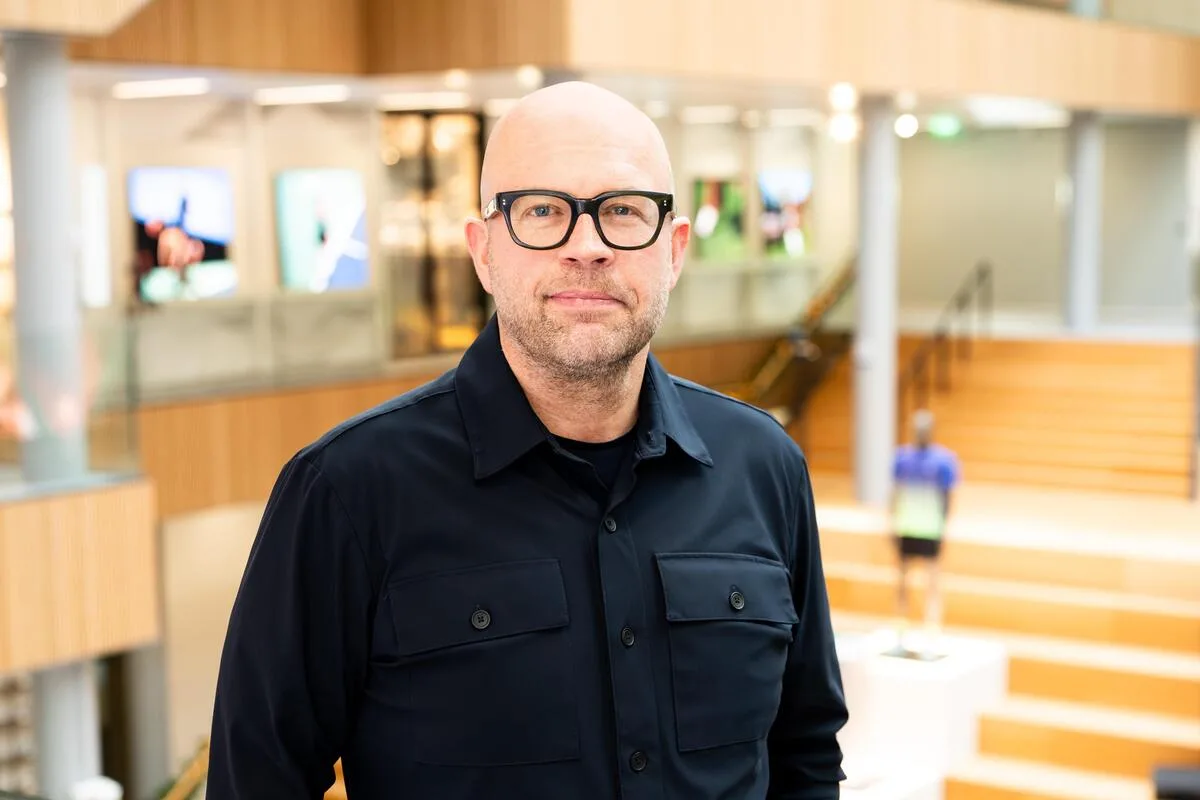 Wyniki finansowe ASICS EMEA 2025. Na zdjęciu: Carsten Unbehaun, CEO ASICS EMEA. Mężczyzna bez włosów w ciemnych okularach i koszuli w ciemnym kolorze stoi w nowoczesnym, widnym wnętrzu z drewnianymi schodami i przeszklonymi powierzchniami w tle.