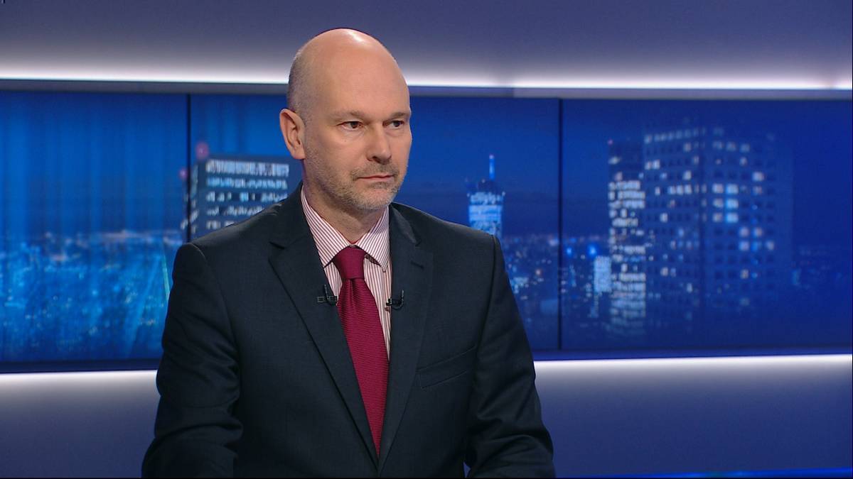 Mężczyzna w garniturze i krawacie siedzi na tle ekran&oacute;w z panoramą miasta nocą.