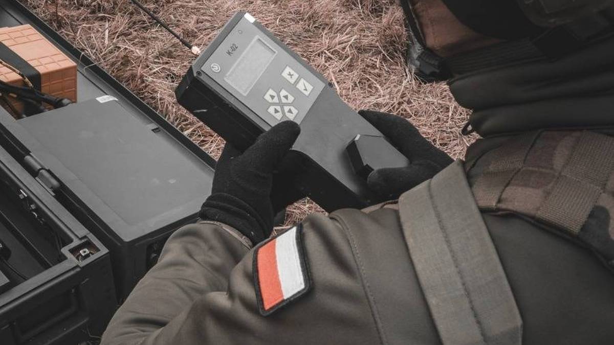 Fragment munduru żołnierza z naszywką w barwach flagi Polski, obok leży sprzęt wojskowy, w tym radio.