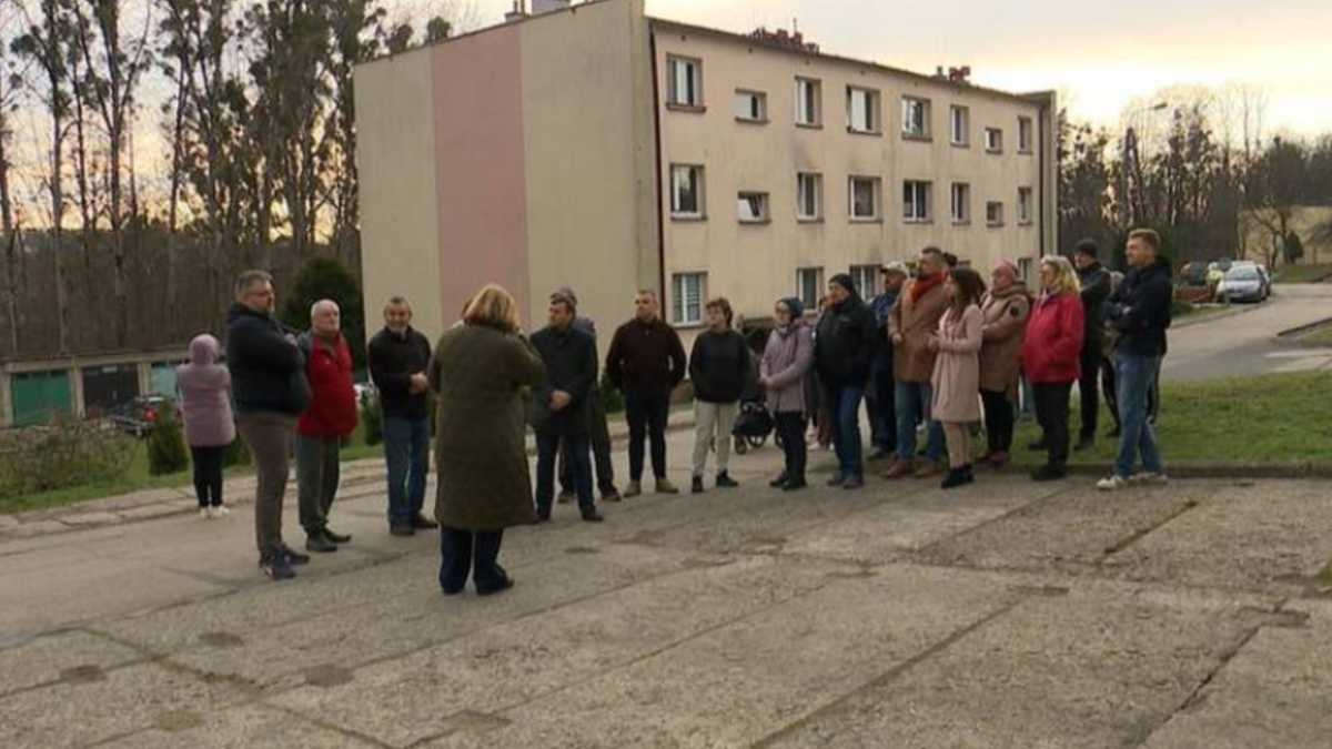 Grupa mieszkańc&oacute;w stoi przed kilkupiętrowym blokiem mieszkalnym, rozmawiają ze sobą z widocznym zaniepokojeniem. Tło stanowi teren zielony z drzewami.