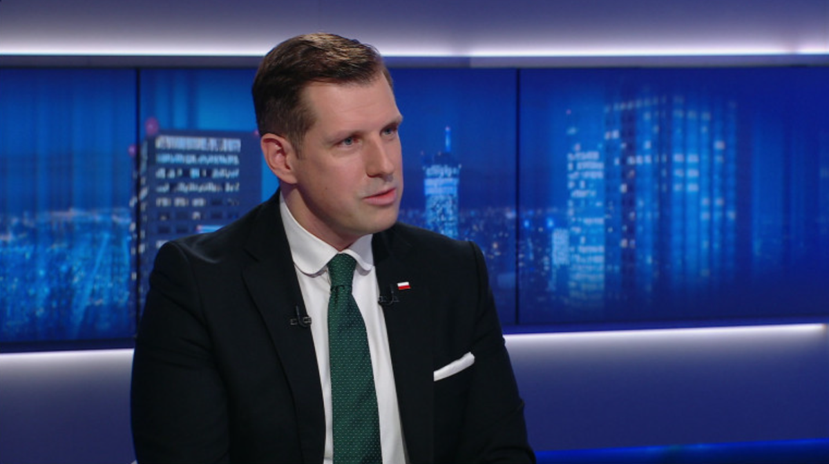 Mężczyzna w garniturze i krawacie siedzi i mówi, w tle widać nocną panoramę miasta.