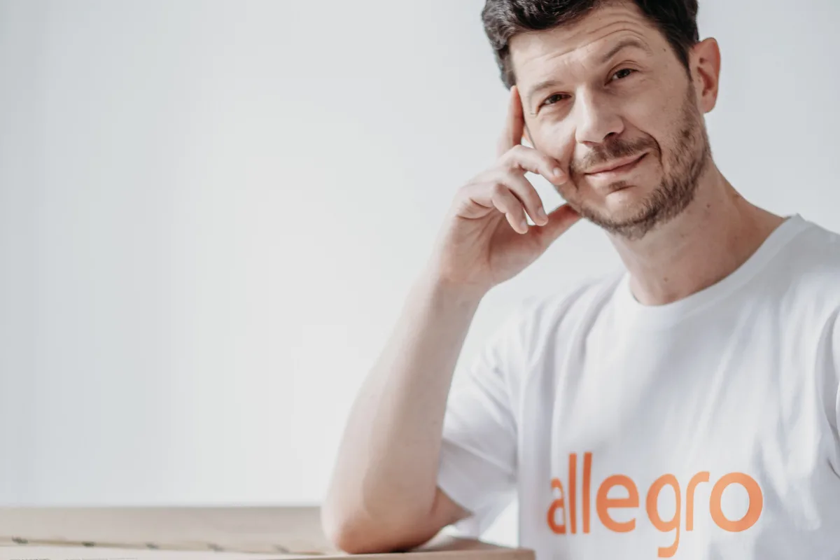 Allegro usługi. Na zdjęciu: Marcin Kuśmierz, prezes Allegro, w koszulce z logo firmy, oparty o kartonowe pudełko z przesyłką.