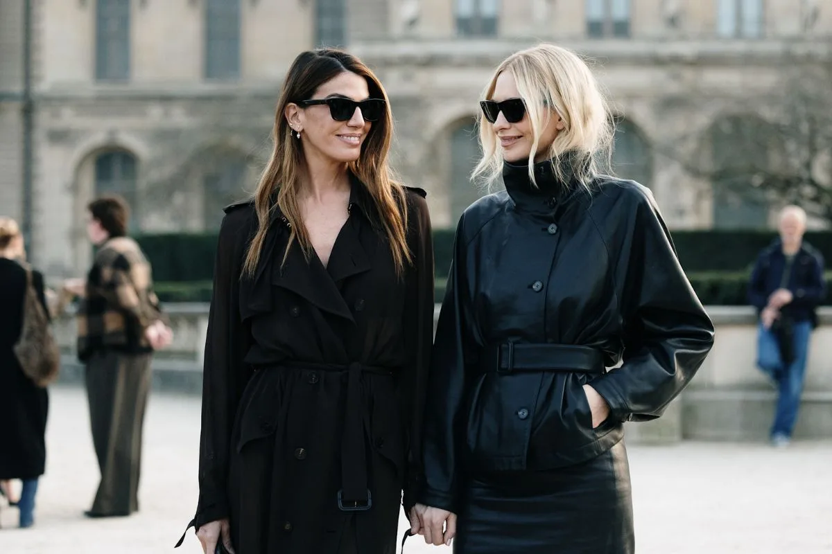 Dwie stylowe kobiety w ciemnych okularach słonecznych przechadzają się na tle klasycznej architektury europejskiej. Jedna nosi czarny, zwiewny płaszcz typu trencz, a druga krótką, skórzaną kurtkę z paskiem w talii i pasującą spódnicę. Te stylizacje odzwierciedlają trendy, które promują czołowe platformy handlowe w Europie w 2026 roku, stawiając na nowoczesny minimalizm oraz materiały wysokiej klasy.