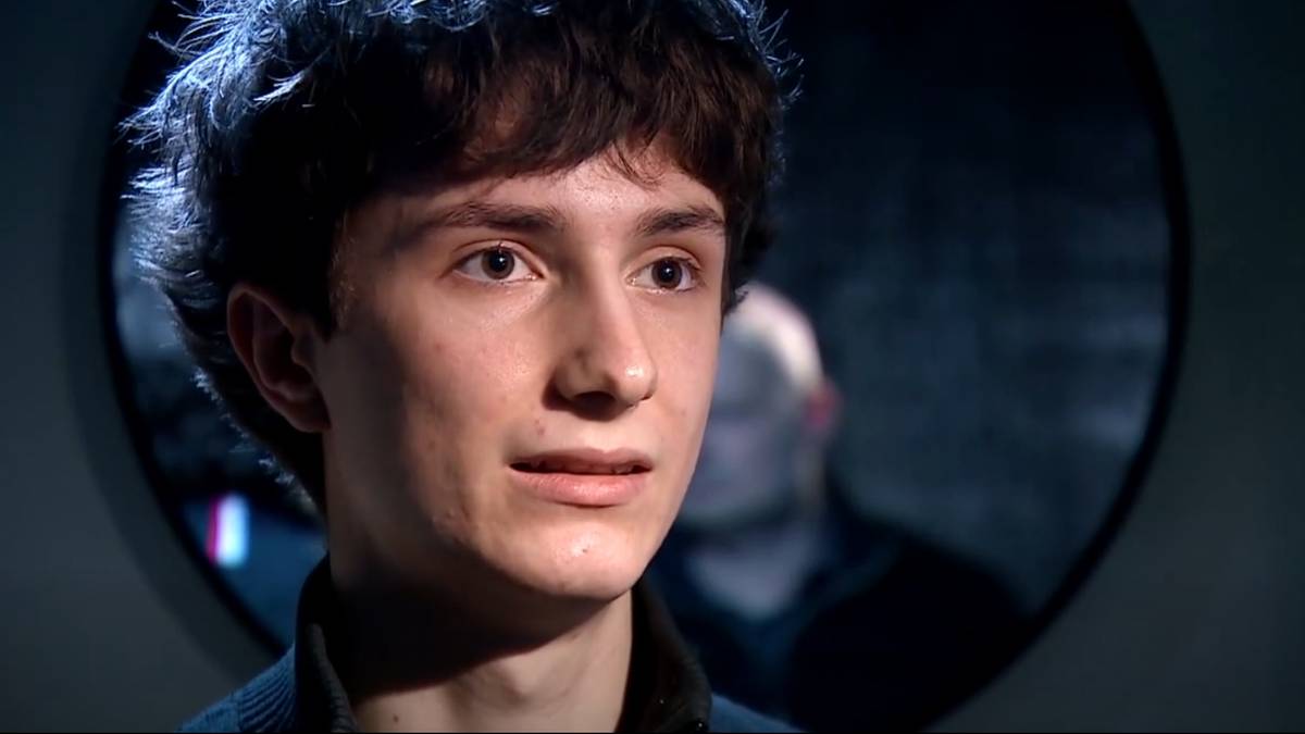 Portret młodego mężczyzny z ciemnymi, kręconymi włosami, skierowany lekko w bok. Na drugim planie, w tle, widoczna jest sylwetka innego mężczyzny w okręgu.