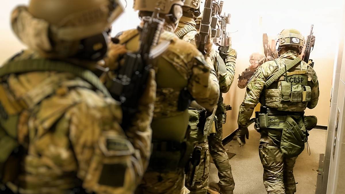 Grupa żołnierzy w pełnym rynsztunku bojowym, każdy uzbrojony w karabin, porusza się w ciasnym korytarzu.