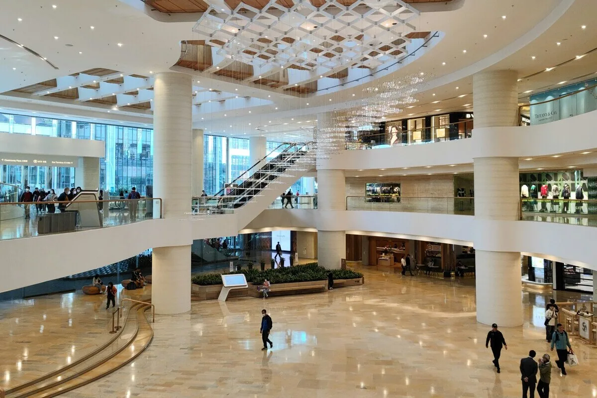 Działka po galerii Plaza w Krakowie. Przestronne, nowoczesne wnętrze wielopoziomowego centrum handlowego z wysokim sufitem, dużymi białymi kolumnami, ruchomymi schodami oraz geometryczną instalacją świetlną zawieszoną nad otwartym atrium; na marmurowej posadzce i balkonach galerii widać spacerujących ludzi oraz witryny sklepów.