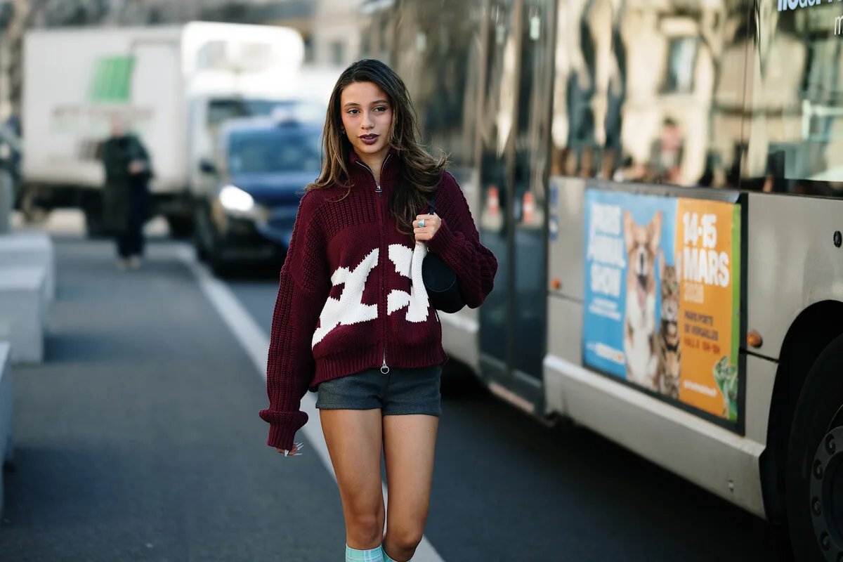 Opóźnienia dostaw fast fashion, Zara, H&amp;M, Primark. Modelka w bordowym swetrze z napisem Zara spacerująca ulicą miasta obok autobusu – przykład stylu street style i odzieży z branży fast fashion.