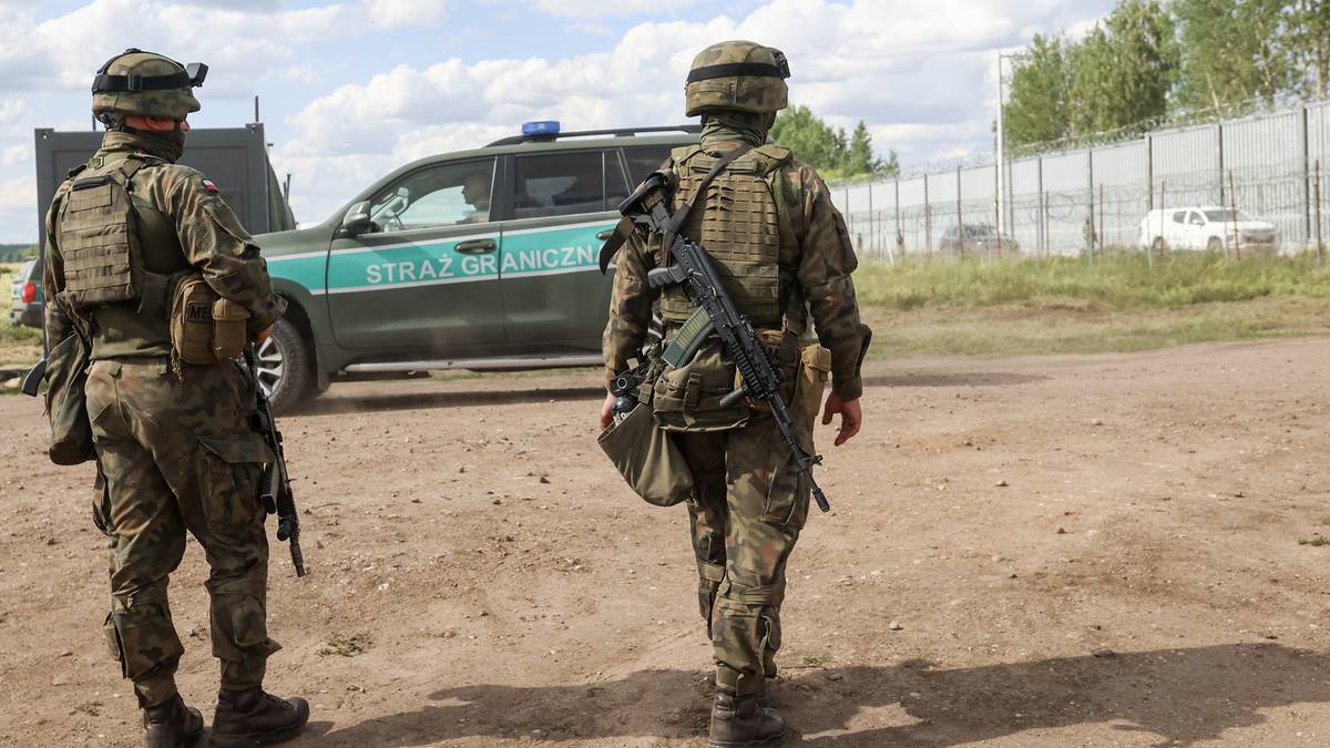 Dw&oacute;ch żołnierzy w pełnym rynsztunku wojskowym stoi na ziemi, a za nimi widać samoch&oacute;d służbowy oraz ogrodzenie graniczne.