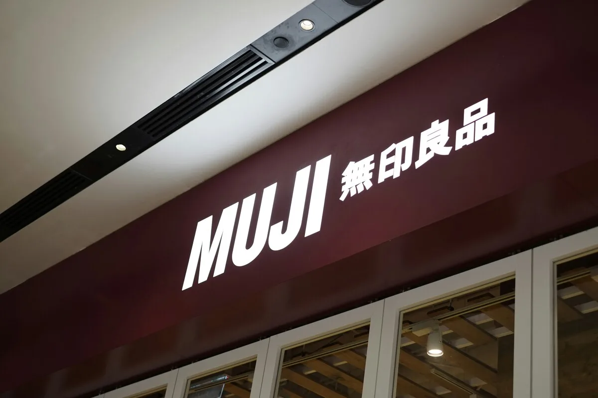 Ekspansja Muji w Europie. Zdjęcie przedstawia szyld sklepu MUJI ulokowany nad wejściem do placówki w centrum handlowym. Na bordowym tle widnieje okazały, biały napis „MUJI”, a tuż obok niego umieszczone są japońskie symbole kanji. Poniżej szyldu dostrzec można oszkloną witrynę oraz fragment wnętrza sklepu z elementami drewnianego wystroju i oświetleniem sufitowym. Całość oddana jest w minimalistycznym, nowoczesnym stylu, typowym dla tej marki.