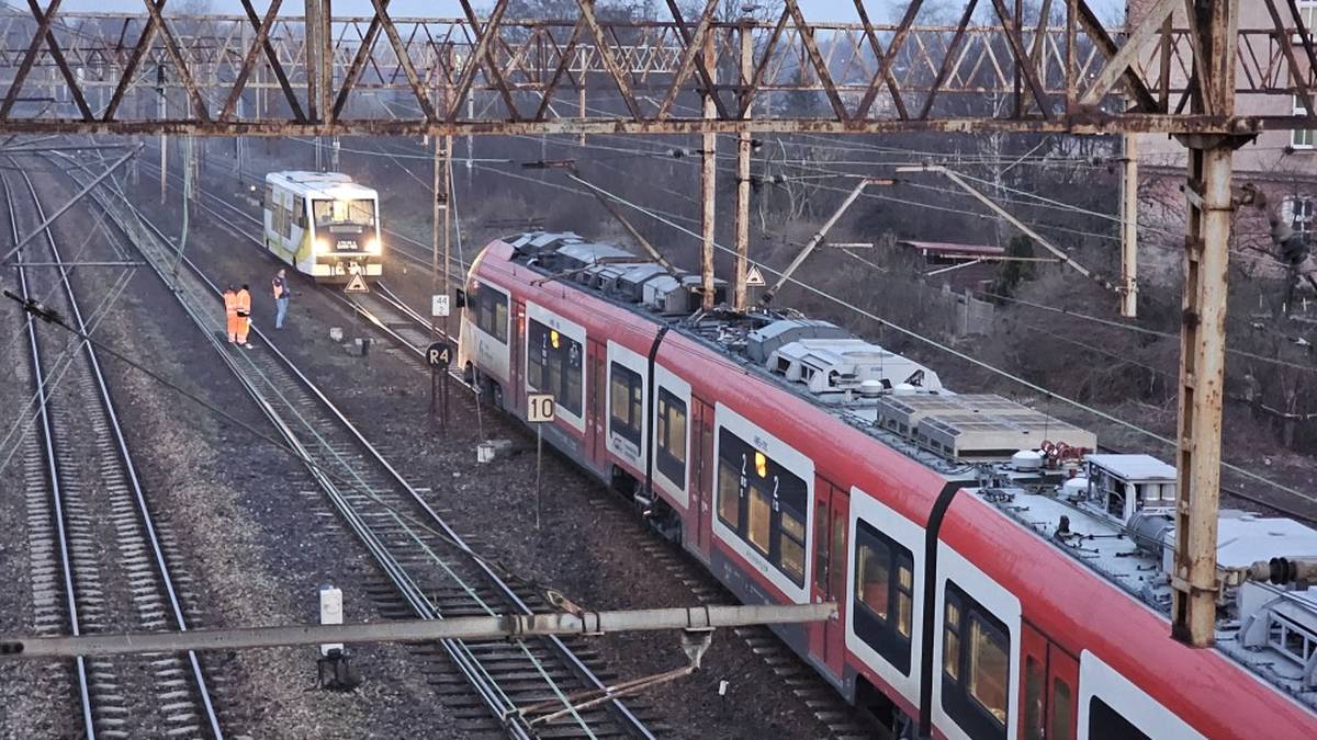 Dwa pociągi jadące tym samym torem kolejowym w Gnieźnie, jeden czerwono-biały, drugi biały, z widoczną infrastrukturą kolejową.