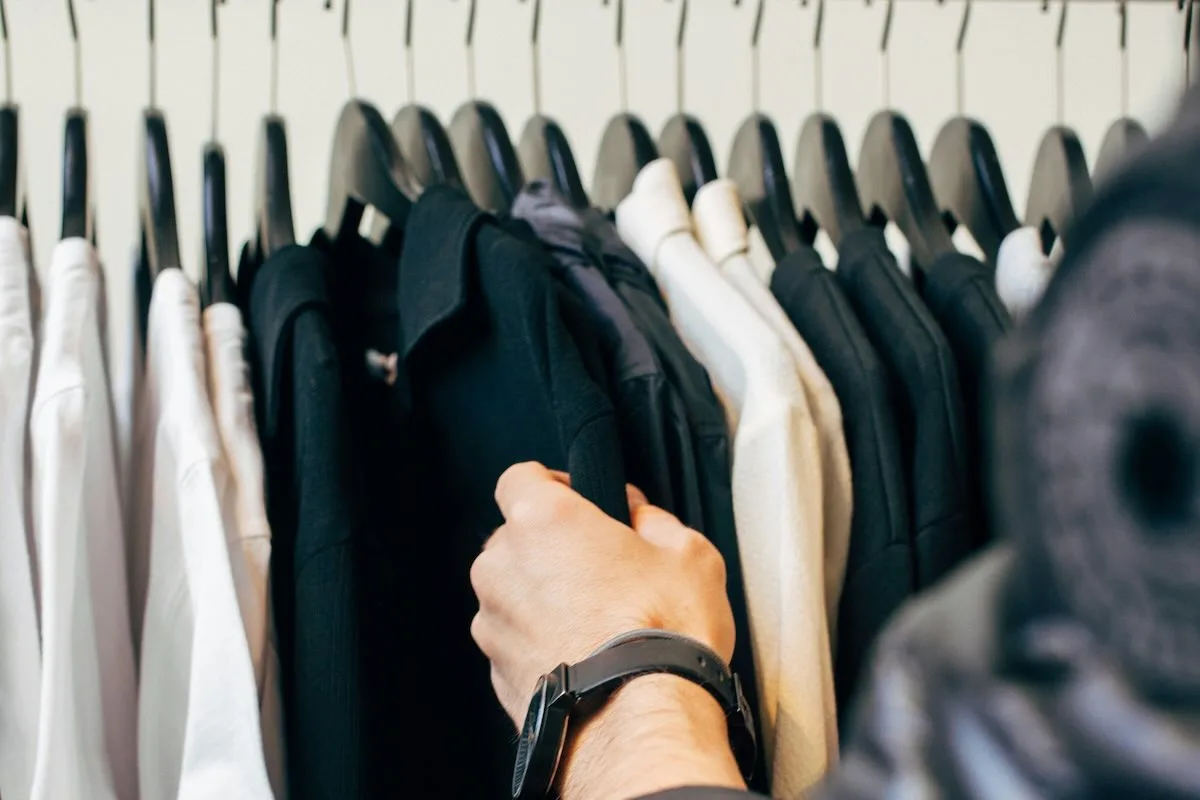 Czy centra handlowe są otwarte w Wielki piątek 2026? Na zdjęciu osoba przeglądająca ubrania na wieszakach w szafie, wybierająca czarną koszulę spośród innych ubrań w stonowanych kolorach.