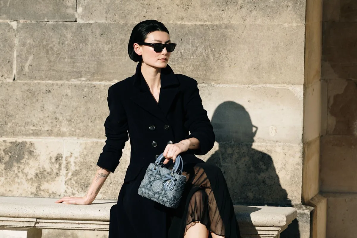 Kobieta w eleganckim, czarnym płaszczu i ciemnych okularach siedzi na kamiennej ławce, trzymając luksusową torebkę. Stylizacja idealna na formalne okazje i wiosenne spacery, gdy zastanawiamy się, czy Wielki Piątek 2026 to dzień wolny od pracy i jak zaplanować świąteczny czas w dobrym stylu.