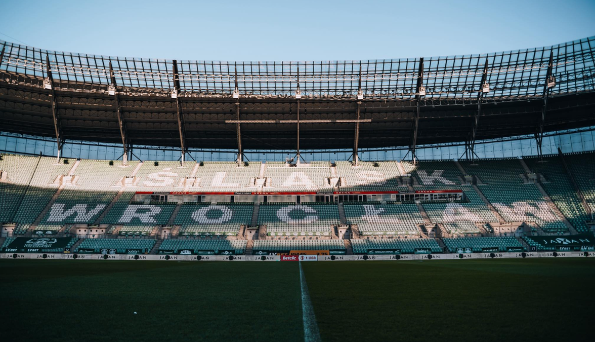 Widok na trybuny stadionu z napisem "ŚLĄSK WROCŁAW" widocznym na zielonych siedzeniach oraz na murawę boiska.