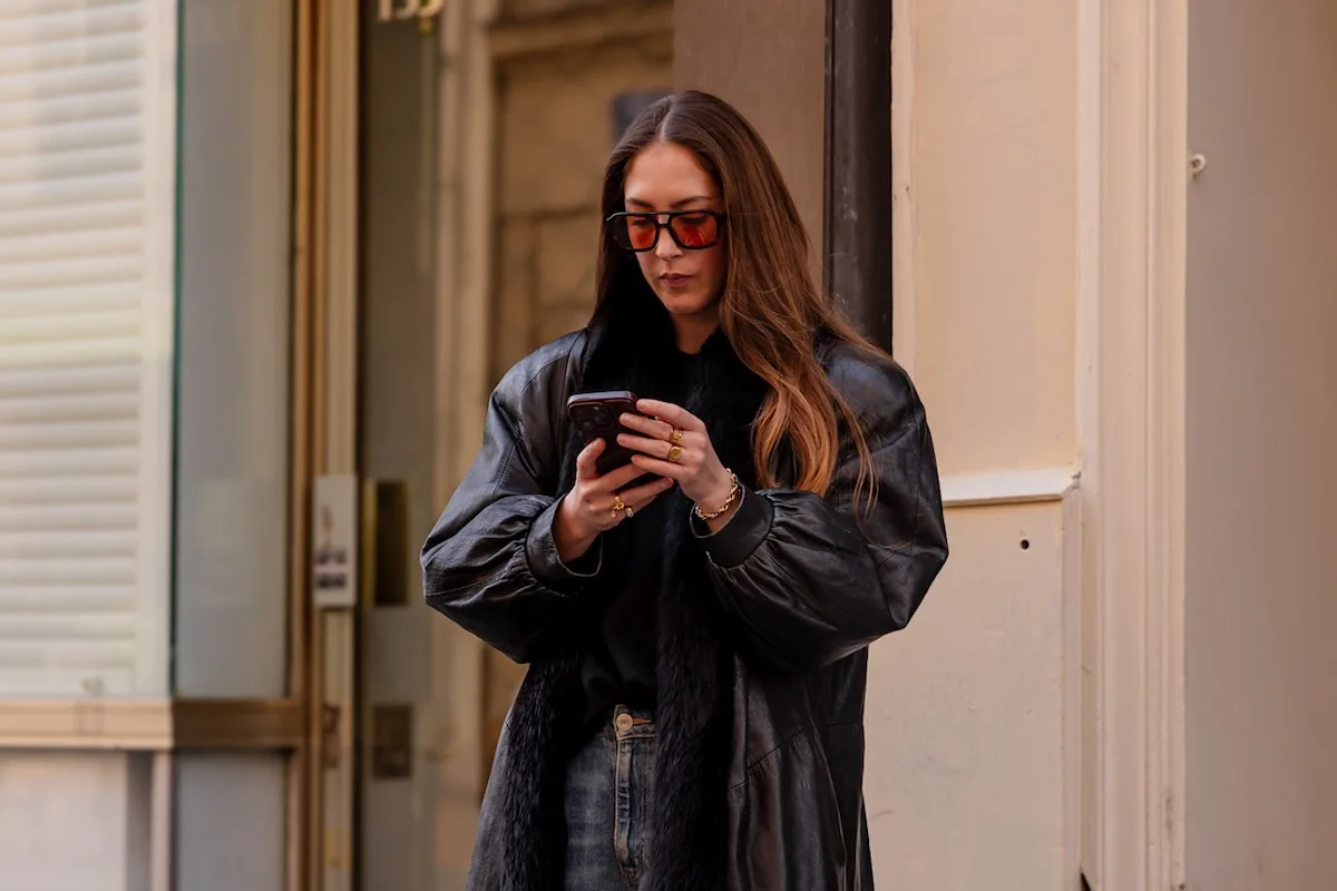 Technologiczne tendencje, które przeobrażą sektor mody w 2026. Kobieta odziana w czarny skórzany płaszcz i obszerne okulary słoneczne stoi na ulicy i używa smartfona.