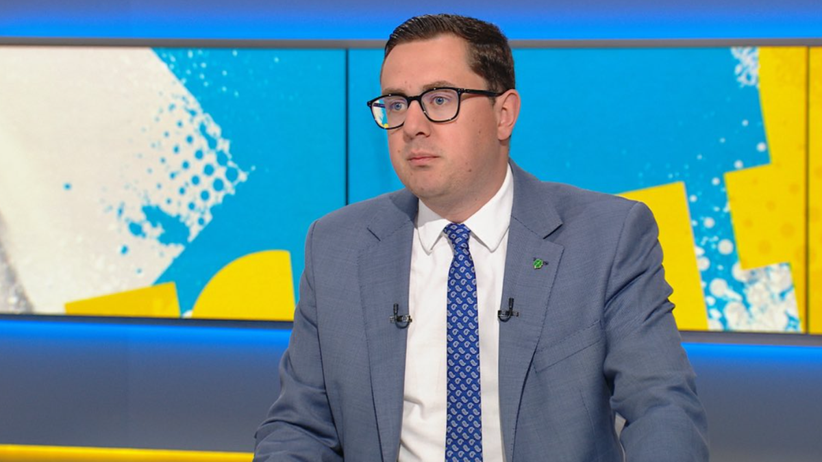 Mężczyzna w niebieskim garniturze i okularach siedzi przed kolorowym tłem studia telewizyjnego.