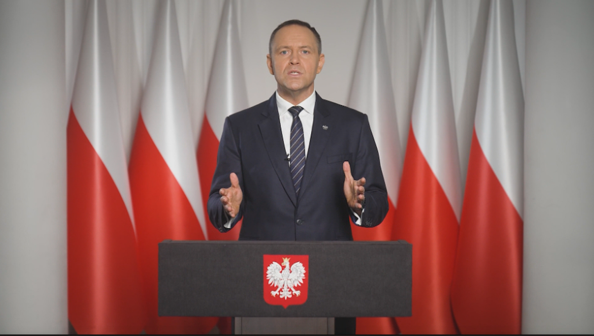 Prezydent Karol Nawrockie przemawia stojąc za m&oacute;wnicą na tle flag Polski.