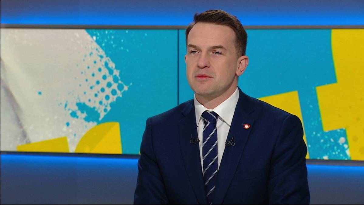 Mężczyzna w granatowym garniturze i pasiastym krawacie siedzi przed kolorowym tłem studyjnym.