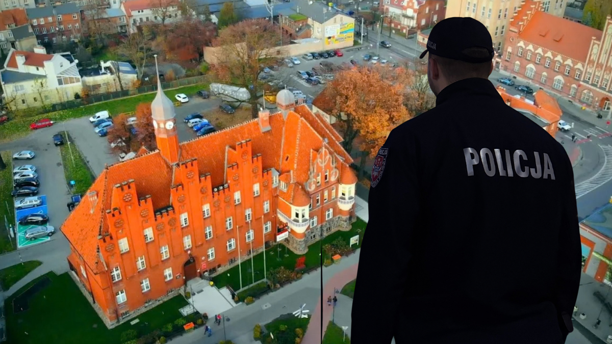 Policjant w mundurze z napisem "POLICJA" na plecach stoi tyłem do widza i obserwuje budynek urzędu miasta.