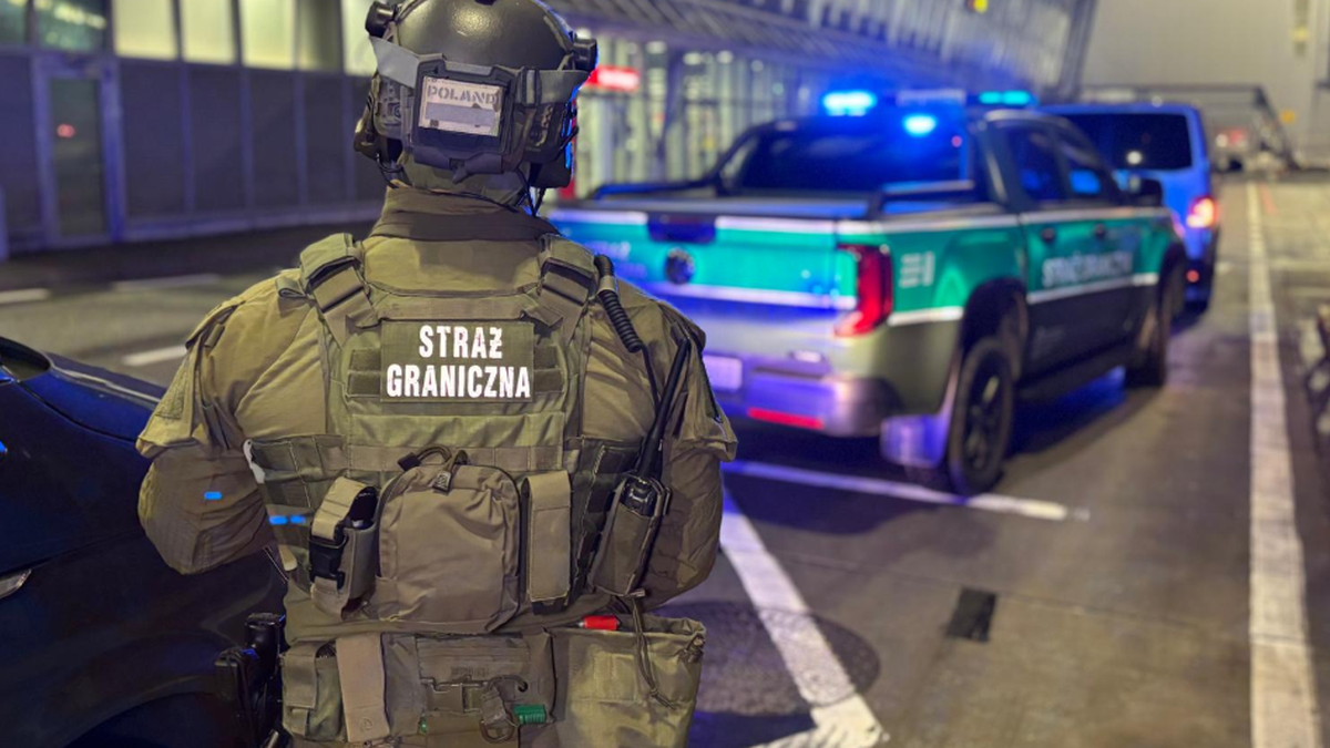 Funkcjonariusz Straży Granicznej w umundurowaniu taktycznym stoi tyłem, obok radiowozu z włączonymi niebieskimi światłami.