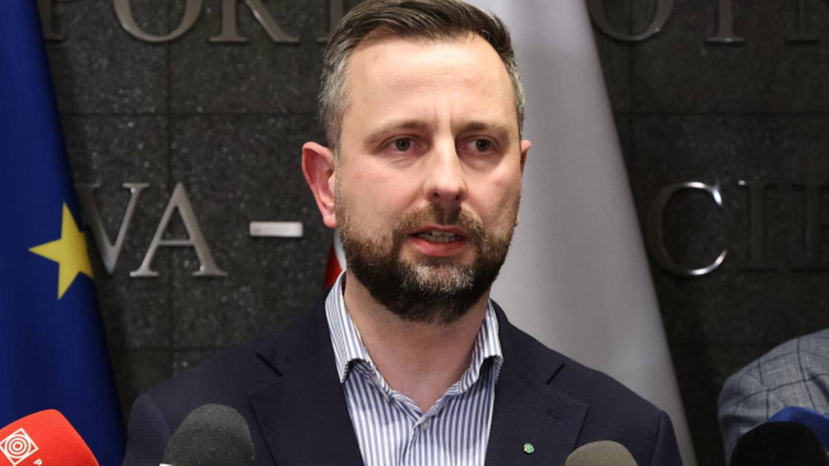 Mężczyzna z brodą, ubrany w koszulę w paski i granatową marynarkę, przemawia przed grupą mikrofon&oacute;w.