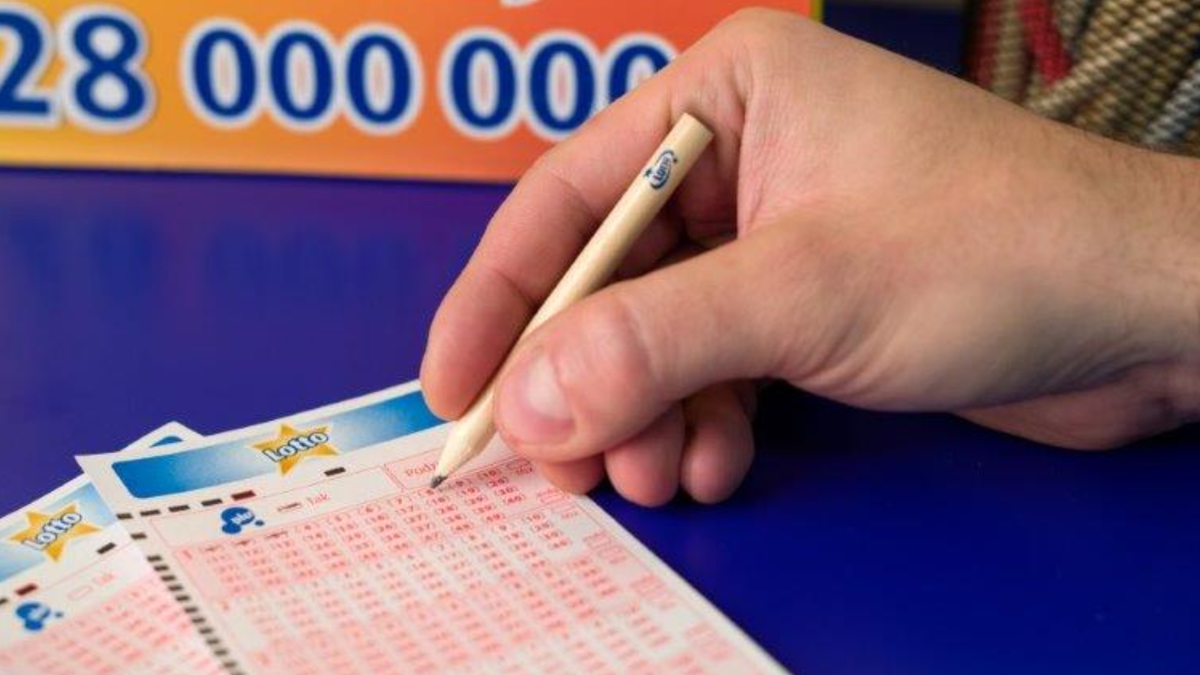 Dłoń wypełniająca kupon Lotto oł&oacute;wkiem, z widocznym fragmentem planszy z kwotą kumulacji.