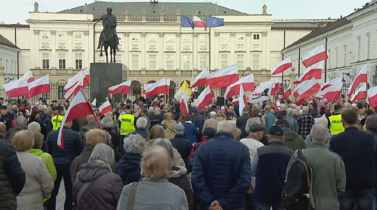 Tłum ludzi trzymających polskie flagi na placu przed Pałacem Prezydenckim w Warszawie. W tle widoczny pomnik konny.