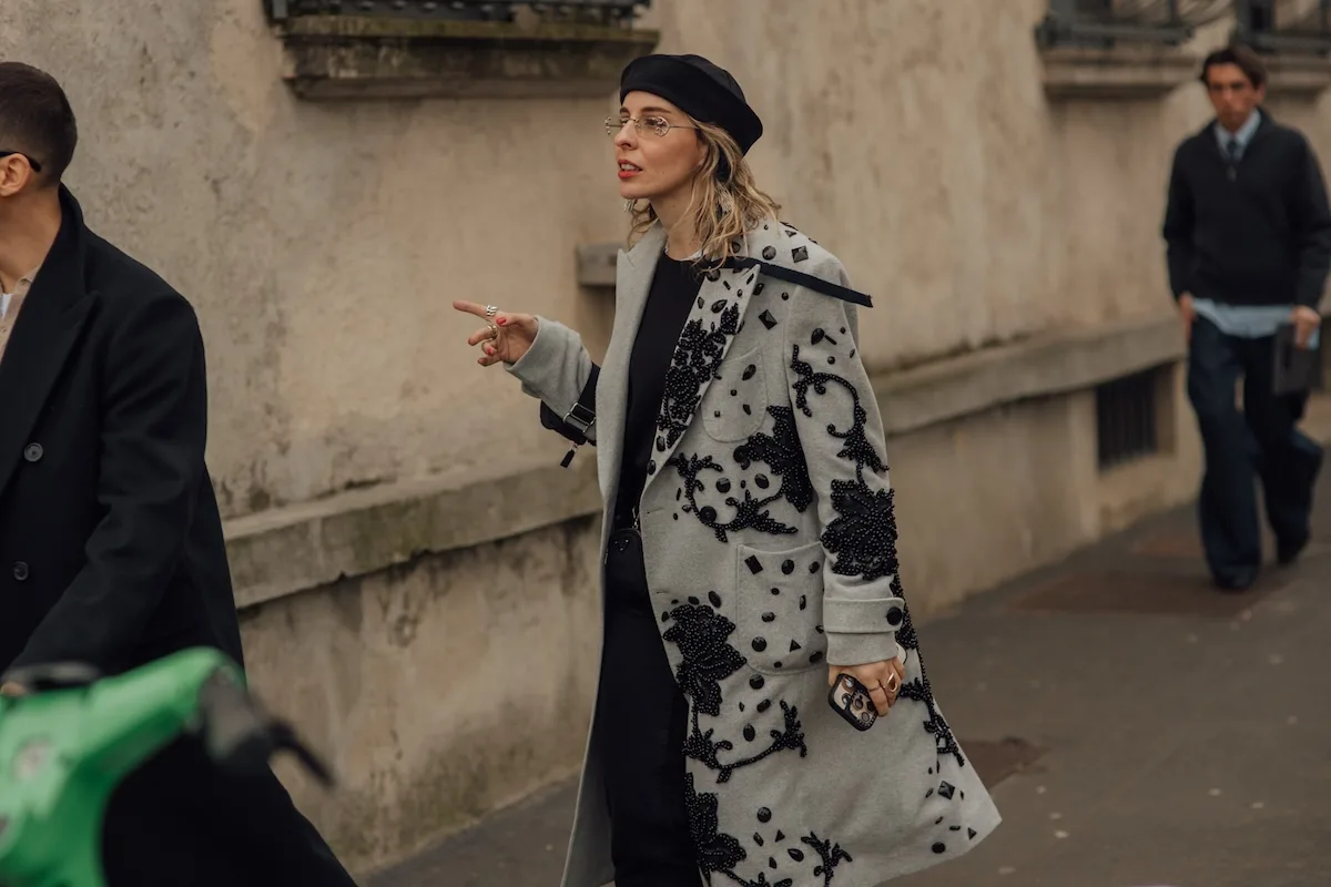 Kobieta w eleganckim okryciu wierzchnim i berecie kroczy ulicą i wskazuje dłonią – miejska scena street style, fotografia ilustracyjna.