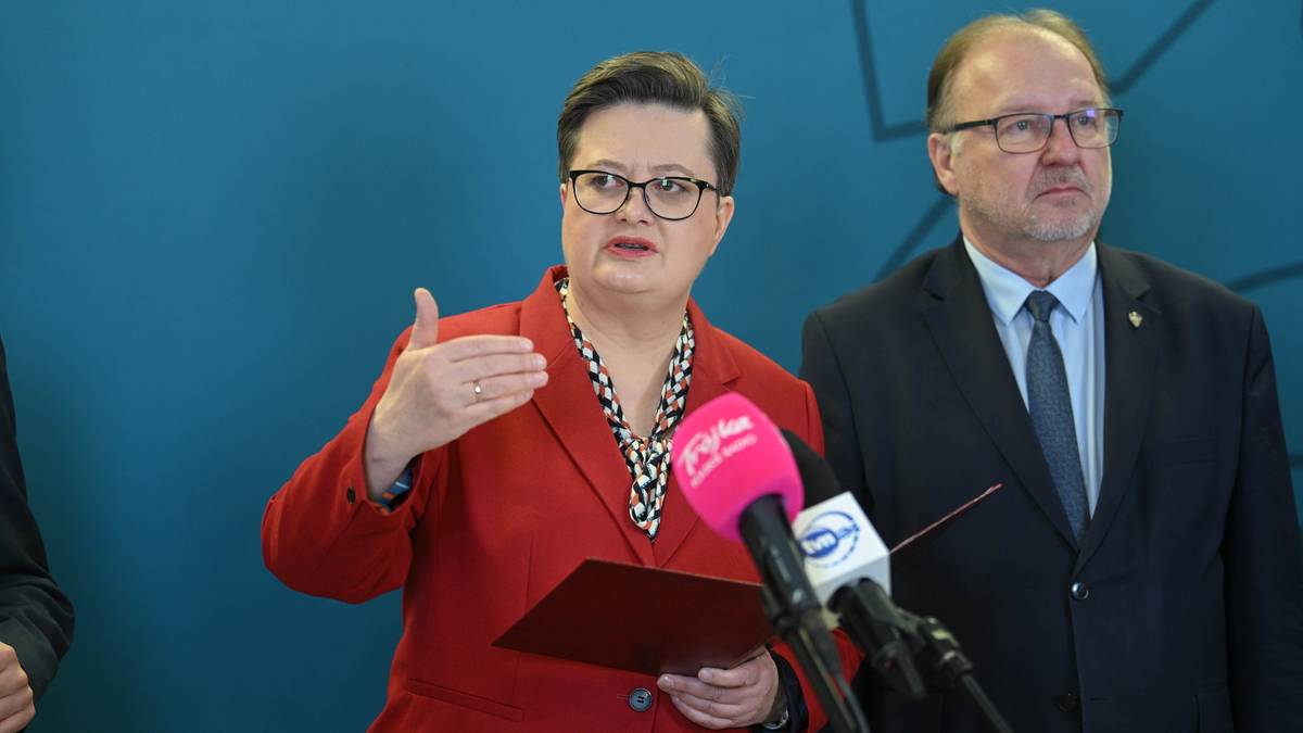 Kobieta w czerwonym żakiecie gestykuluje, trzymając w ręku teczkę i mikrofon. Obok stoi mężczyzna w garniturze i okularach.