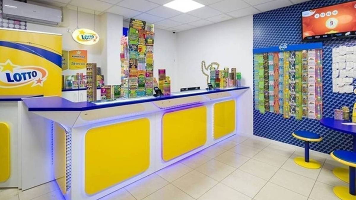 Widok wnętrza punktu sprzedaży gier liczbowych Lotto, z ladą sprzedażową, ekspozycją produkt&oacute;w i ekranem informacyjnym.